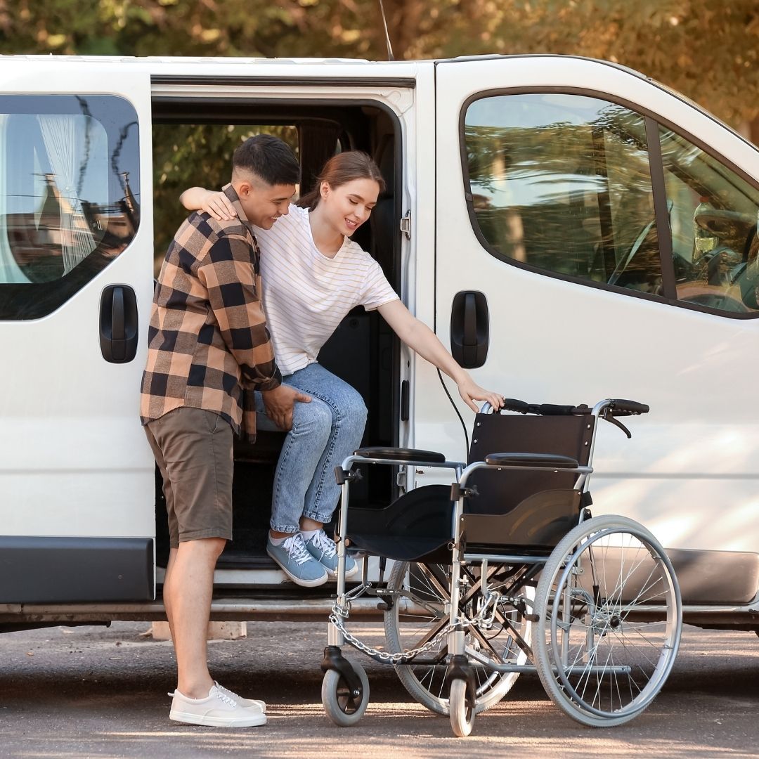 A carer helping a passenger step out of a white accessible van, with a wheelchair ready beside them.
